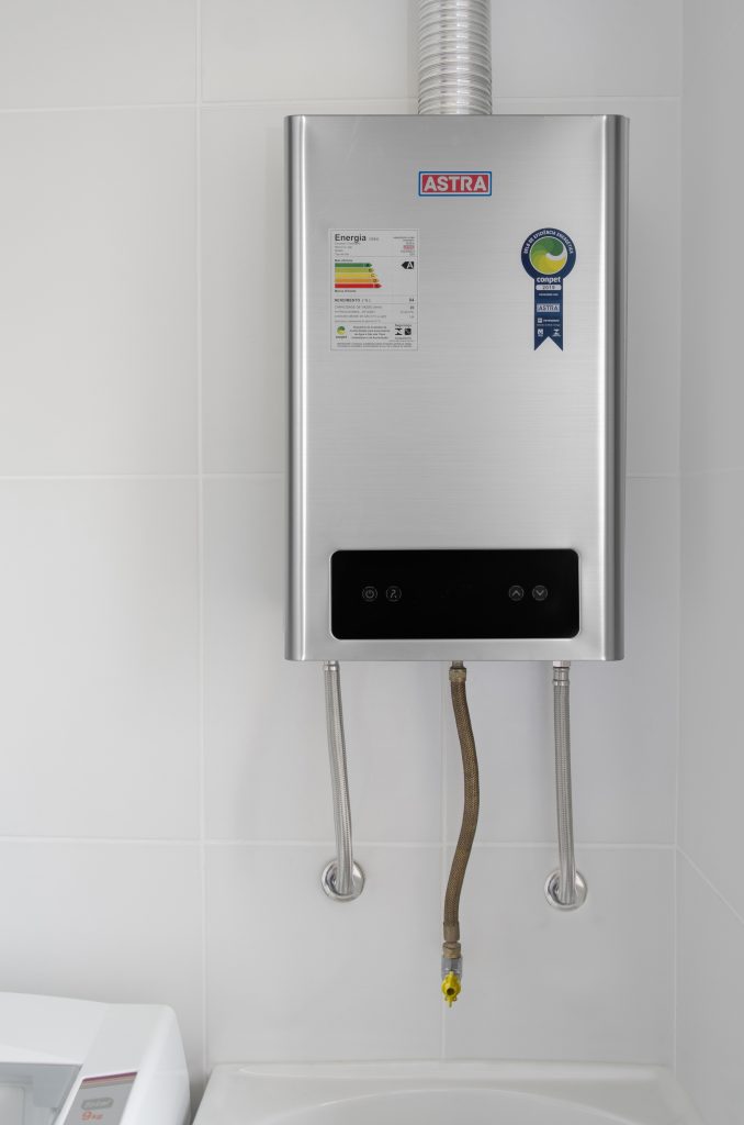 A imagem mostra um aquecedor de água a gás cromado, com ajuste digital, instalado em uma parede de cor branca sólida.