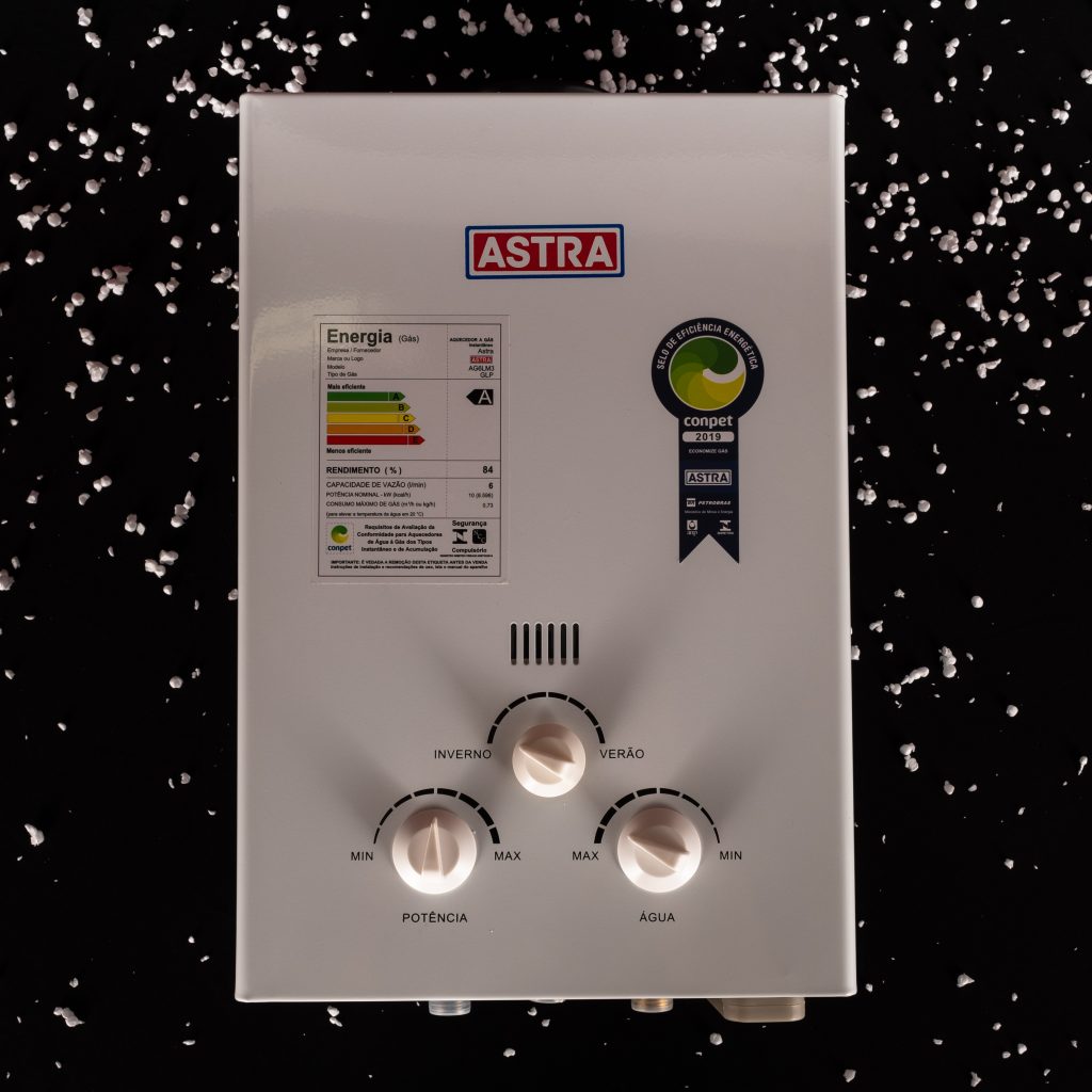 A imagem mostra um aquecedor a gás branco da Astra. Ele possui três ajustes manuais, de potência, água e de temperatura. O fundo da imagem é preto e é possível ver flocos branquinhos simulando flocos de neve, para representar o frio.