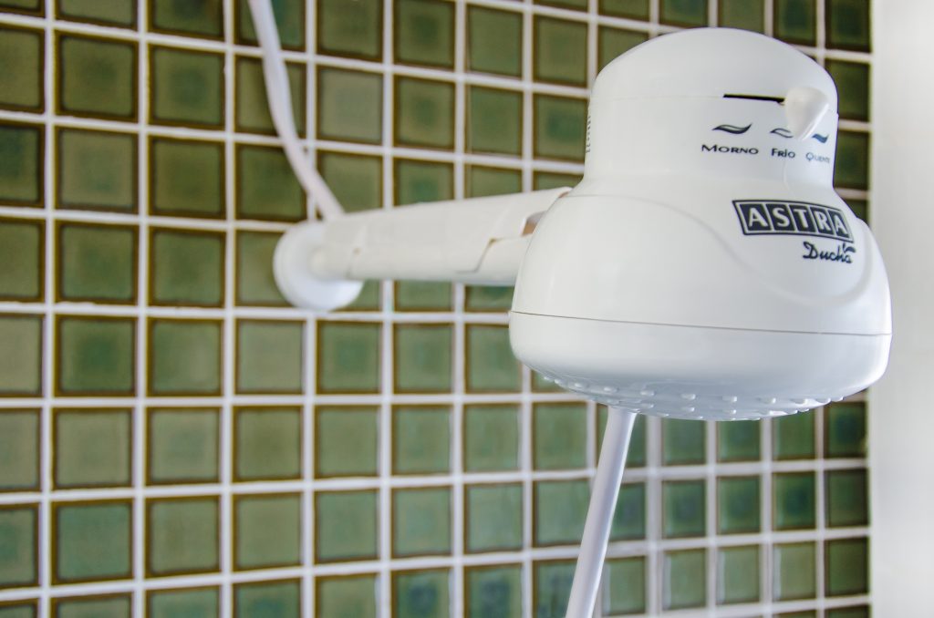 A imagem mostra um um dos tipos de chuveiro elétrico simples da Astra, com três temperaturas, instalado em um banheiro com revestimento verde quadriculado.