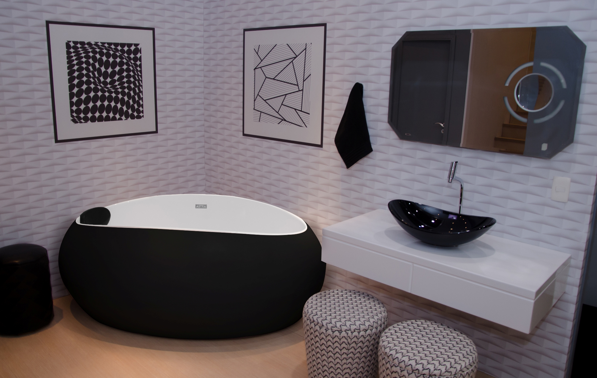 A imagem mostra um banheiro com uma banheira oval preta, em material que a deixa fosca. A banheira está instalada no canto entre duas paredes. Ao lado direito dela, há uma cuba preta, instalada em um gabinete branco.