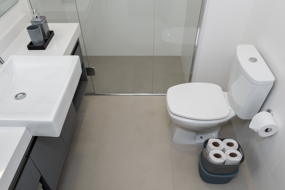 A imagem mostra um banheiro, com vaso sanitário e caixa acoplada brancos, caixas plásticas que guardam rolos de papel higiênico e um suporte de papel higiênico instalado na parede. Também é possível ver uma cuba sob um gabinete.