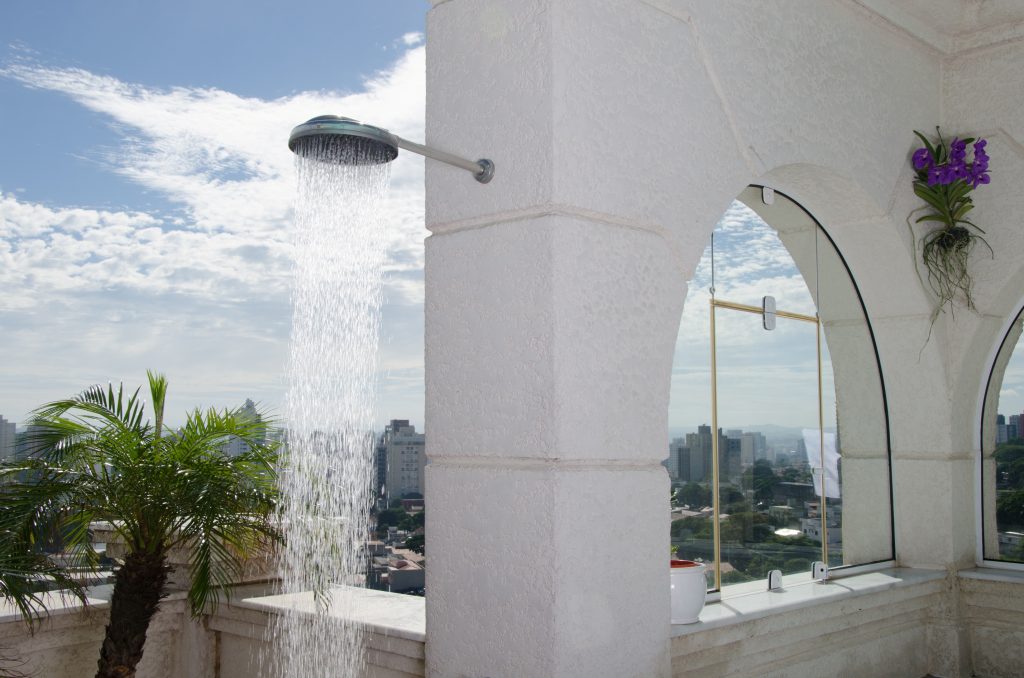 A imagem mostra um chuveiro frio instalado na área externa de uma casa. Ao fundo, é possível ver alguns prédios da vizinhança.