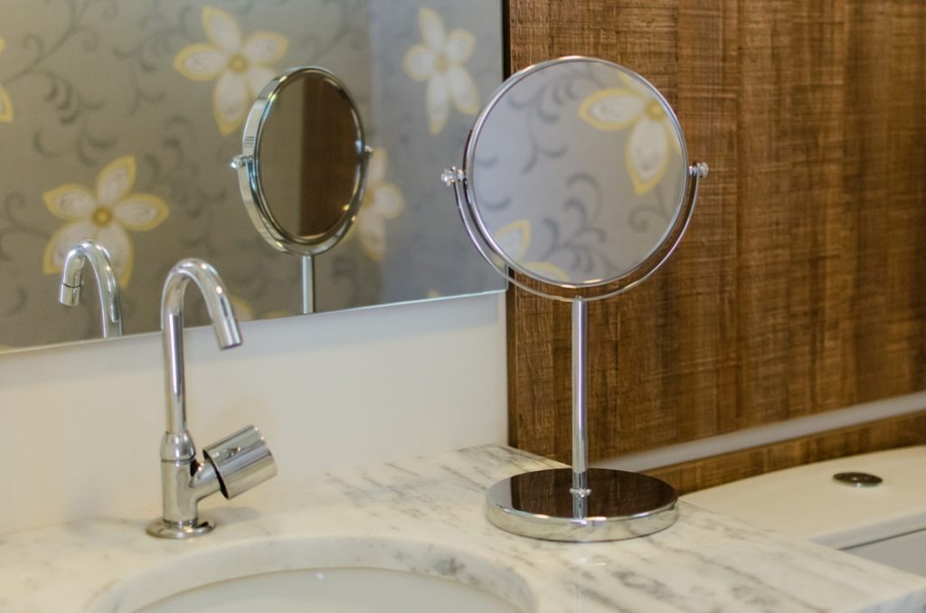 Um espelho redondo pequeno, está em cima de uma pia de banheiro. Ao fundo há uma placa de madeira na parede.