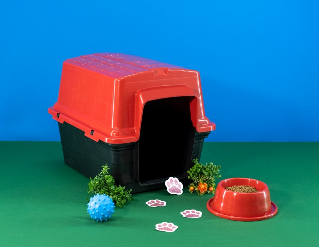 A imagem mostra uma caixa de transporte para cachorro com a tampa vermelha e a parte de baixo preta, uma bolinha de plástico na cor azul e um comedouro para cachorro anti formiga, que é vermelho. Todos os produtos são produzidos pela Astra.
