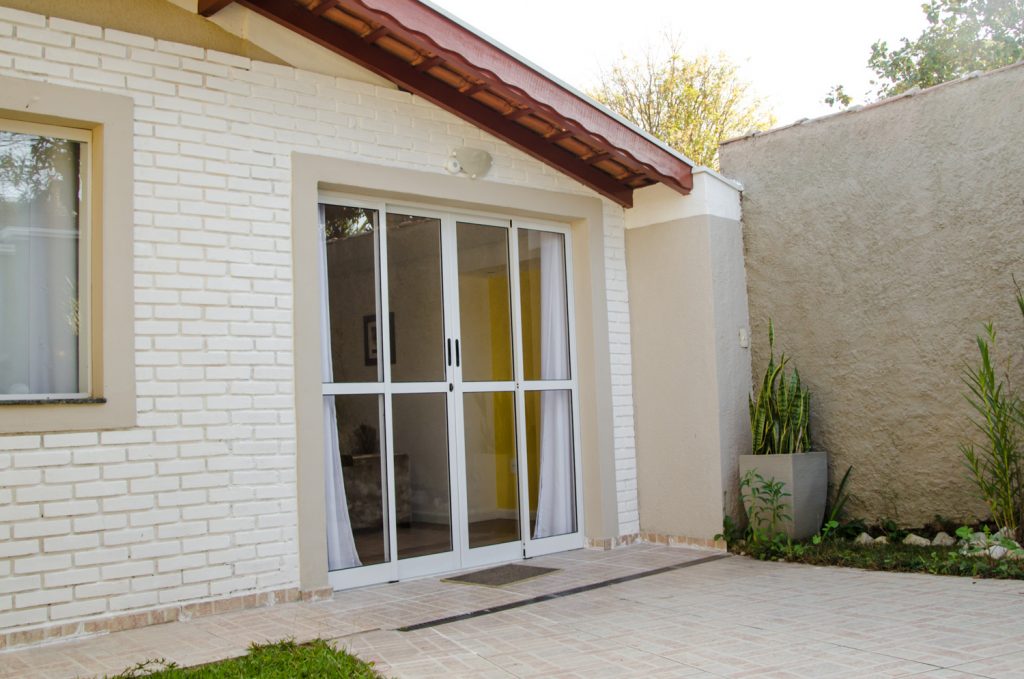 A imagem mostra uma porta de alumínio de correr vista da parte de fora da casa. A porta possui vidros em todas as partes e a estrutura de alumínio é branca. Dentro da casa, é possível ver uma cortina branca aberta.