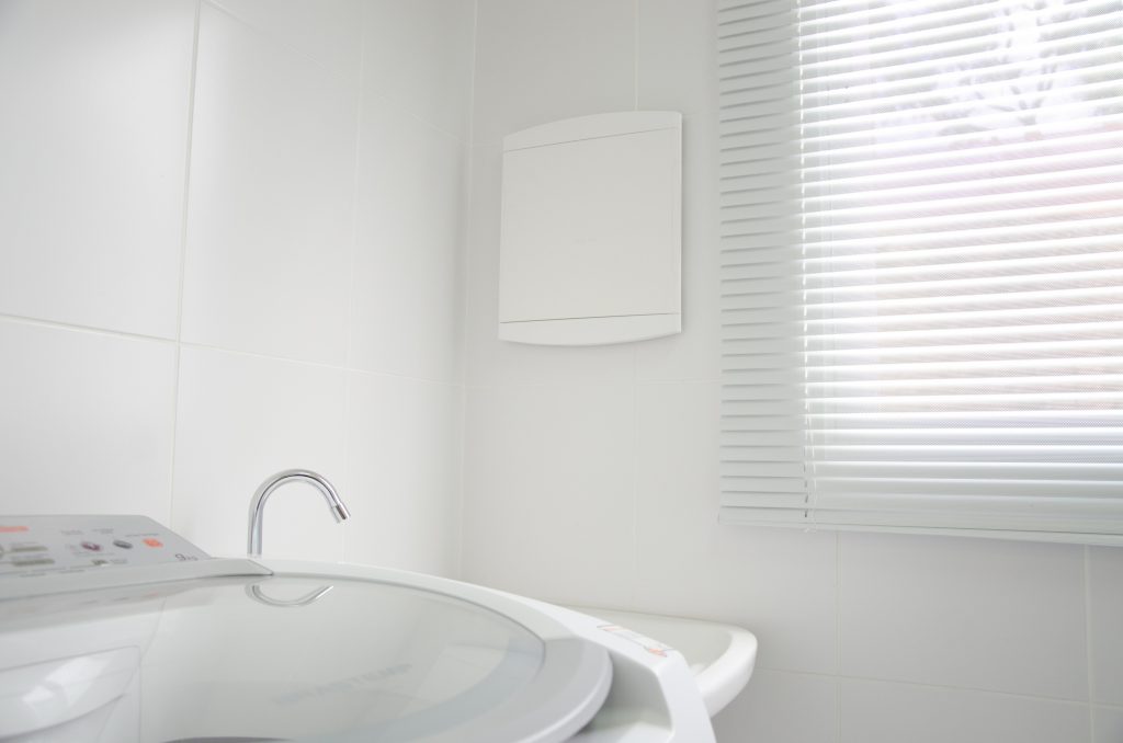 A imagem mostra um quadro de disjuntor instalado na área de serviço de uma casa. Nela, é possível ver um pedaço de uma máquina de lavar, um tanque e uma persiana branca.