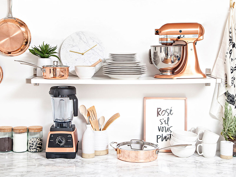 Bancada de cozinha em granito claro e com vários aparelhos, louças e potes em tons de branco, rose gold e madeira.