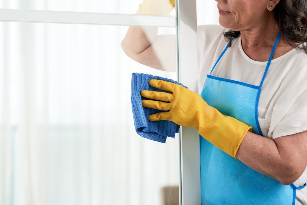 A imagem mostra uma mulher limpando o vidro de uma porta de alumínio com um pano macio. Uma das dicas de como limpar porta de alumínio é com uma mistura de água com detergente neutro, que pode ser espalhada pela porta com uma esponja ou um pano macio, como na imagem.