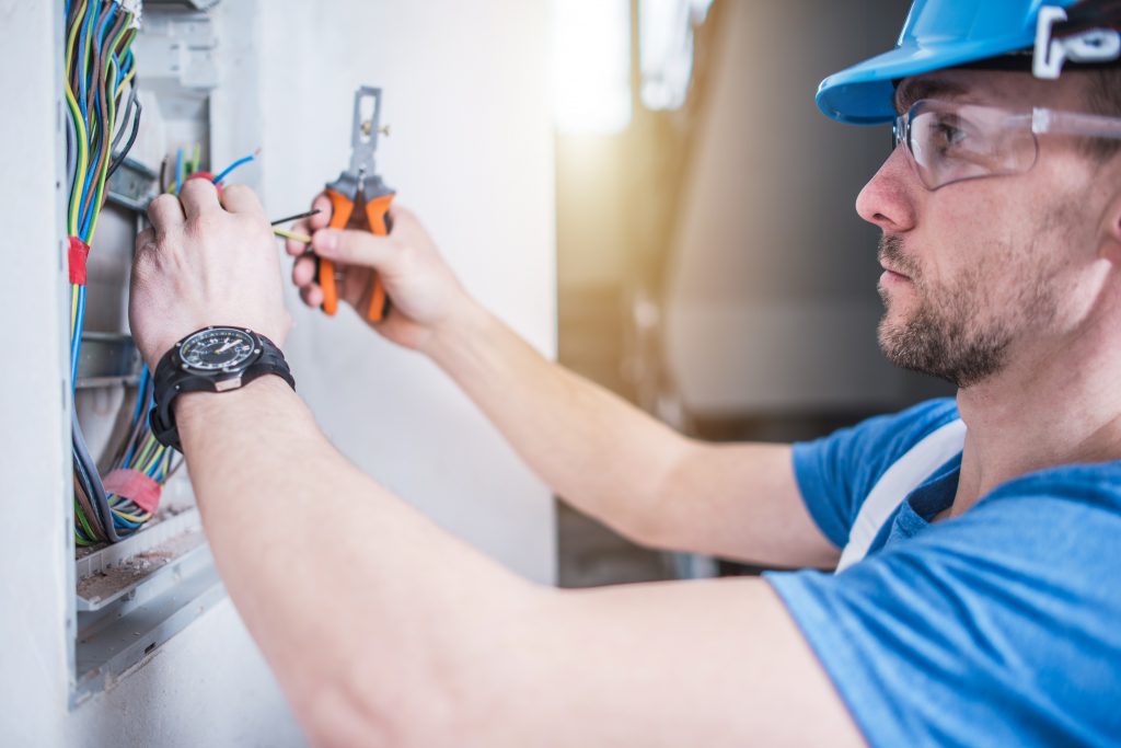 A imagem mostra um eletricista realizando a manutenção elétrica em um quadro de energia. O eletricista é um homem branco e está posicionado no lado direito da imagem, virado de perfil para o quadro de energia, que está no lado esquerdo,