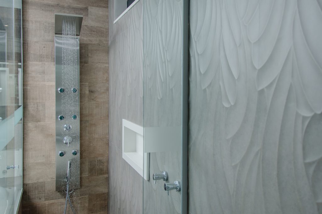 A imagem mostra a área de banho de um banheiro. Na parte central, há um painel de banho cinza instalado em uma parede de aspecto amadeirado. Ao lado direito, a parede possui tom acinzentado e há um nicho embutido de fibra de vidro na cor branca.