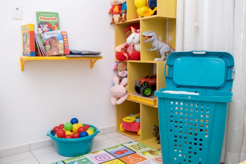 A imagem mostra um espaço do quarto de criança. Nele, há uma prateleira amarela fixada na parede com alguns livros infantis. No chão, abaixo da prateleira, está uma bacia plástica que armazena bolinhas coloridas, como as utilizadas em piscinas de bolinhas. Na parede ao lado, na direita, há uma estante de madeira com brinquedos e ursos de pelúcia. Ao lado dela há um cesto telado azul, que armazena alguns brinquedos.