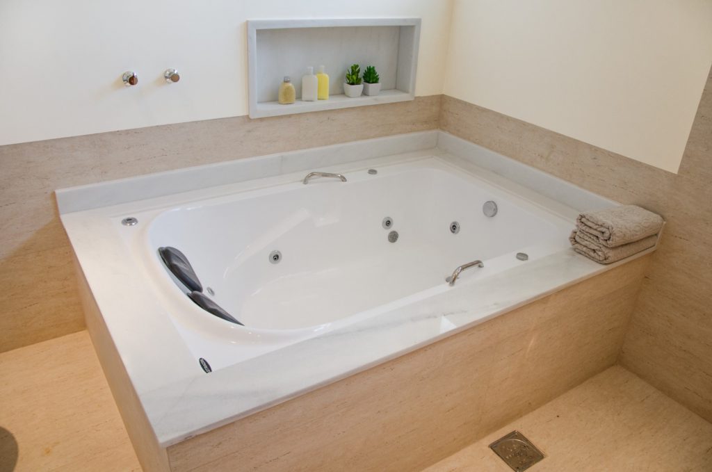 Uma banheira de imersão branca instalada em um banheiro nas cores branco e marrom-claro. Algumas toalhas, alguns produtos de higiene pessoal e dois vasinhos de planta podem ser vistos na imagem.