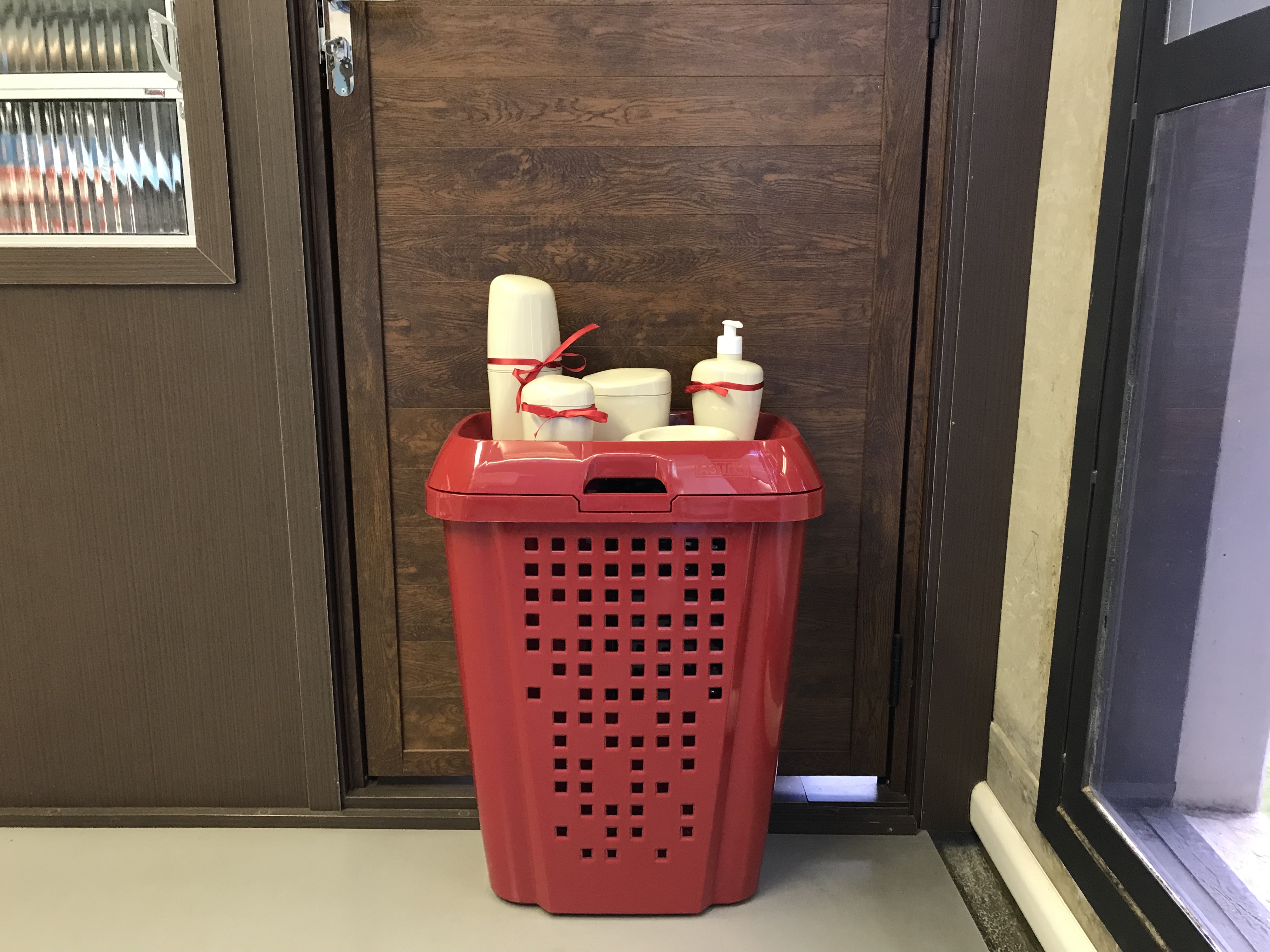 Na imagem um cesto de roupa vermelho aparece na frente de uma porta marrom. Em cima dele há itens de organização de banheiro na cor bege, todos os itens estão envolvidos por um laço vermelho.