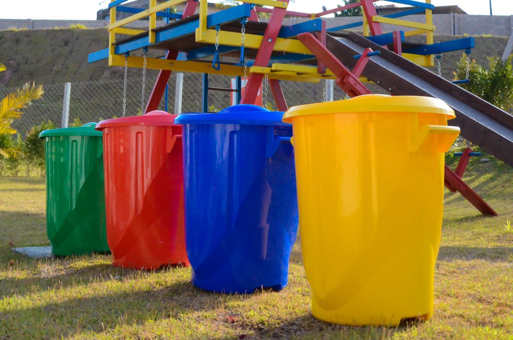 Na foto, é possível ver quatro lixeiras em um jardim, ambas para separar os tipos de reciclagem. Elas estão lado a lado, em uma fila. Da direita para a esquerda, está a lixeira amarela, a azul, vermelha e verde. Ao fundo é possível enxergar um parque infantil.