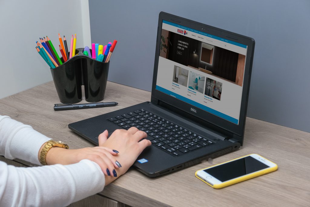 Na foto uma mão mexe em um notebook, a tela de navegação é o site institucional da Astra. O computador está sob uma mesa de madeira clara, junto à um celular de capinha amarela e um porta-objetos que acomoda canetas coloridas. 