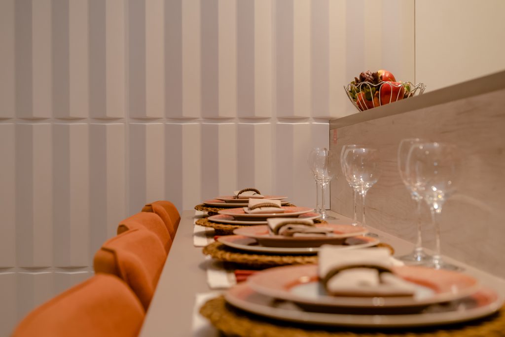 Na foto é possível ver uma parede com o revestimento 3D da Astra, o item possui linhas verticais. Na frente, há uma mesa posta de jantar, com pratos, taças e guardanapos arrumados. Atrás dela há cadeiras laranja. Acima da mesa há uma bancada que acomoda uma fruteira.