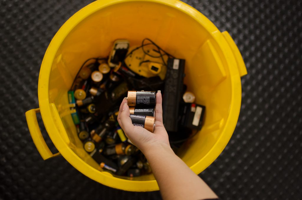 Na imagem é possível ver a mão de uma pessoa branca segurando algumas pilhas em cima da boca de um cesto amarelo. No interior do cesto é possível visualizar alguns cabos e pilhas.