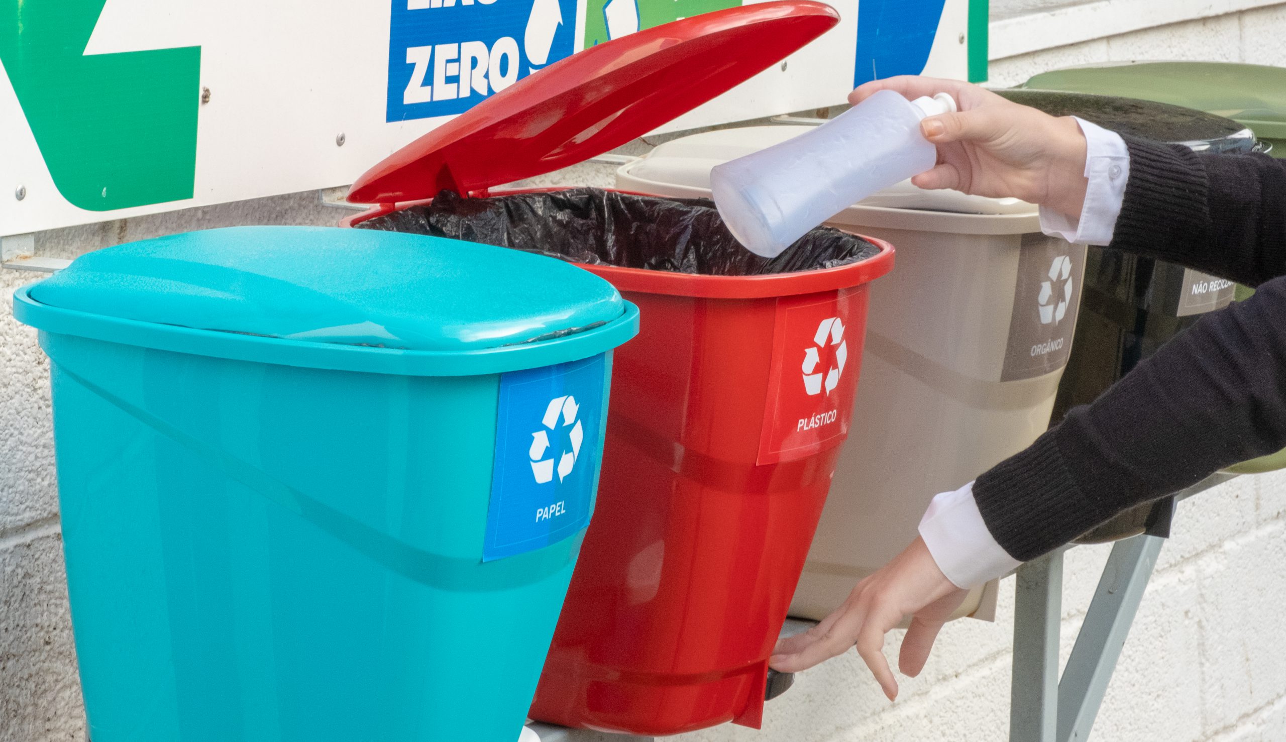 A imagem mostra cinco lixeiras coloridas, utilizadas para descartar corretamente os resíduos reciclados. Da esquerda para a direita, há uma lixeira em tom azul-claro, destinada para o descarte de papel; ao lado dela está uma lixeira vermelha, destinada aos resíduos plásticos, em seguida, há uma lixeira bege, usada para o descarte do lixo orgânico; por fim, há uma lixeira em tom verde-escuro, usada para descartar materiais não reciclados. Também é possível ver as mãos de uma pessoa branca. Com a mão esquerda, ela pressiona a alavanca que abre a lixeira vermelha. A mão direita da pessoa leva uma embalagem plástica ao interior da lixeira.