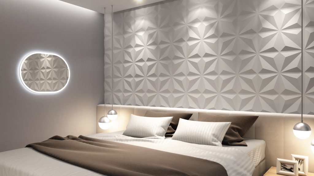 A imagem mostra um quarto. Nele, há uma cama de casal ao centro. Na parede atrás dela há revestimentos 3D brancos, que formam origamis. Também é possível ver um espelho oval com led na parede à esquerda da cama. 