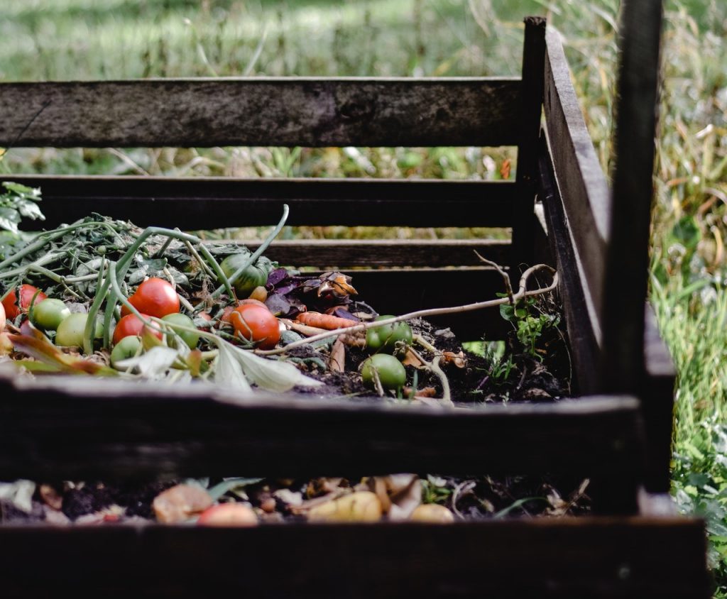 A imagem mostra uma estrutura de madeira colocada sobre o chão. Dentro dela há os restos de frutas e verduras, que são usadas para como fazer a compostagem em casa. 