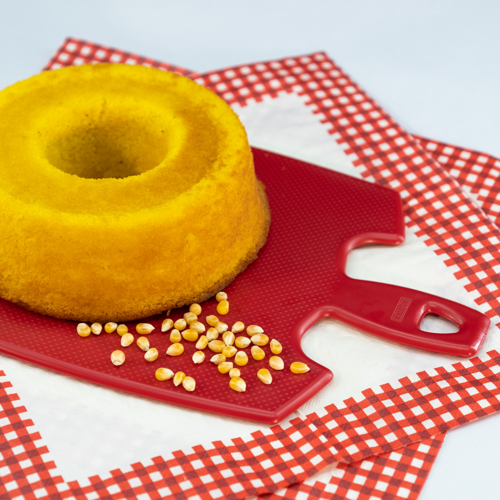 Na foto é possível visualizar um bolo de milho em cima de uma tábua de corte vermelha. Em cima da tábua há grão de milho. A tábua está apoiada em uma toalha brancas com as bordas em xadrez vermelho. 