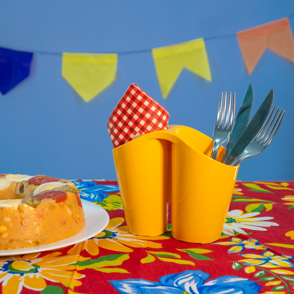 Típico de Festa Junina, o cuscuz está visível na foto em cima de um prato branco. Ao ladoé possível ver um porta-talheres da Astra amarelo que acomoda dois garfos e duas facas, além de alguns guardanapos. Ambos estão em cima de uma toalha vermelha florida. O fundo da foto é roxo com algumas bandeiras típicas. 