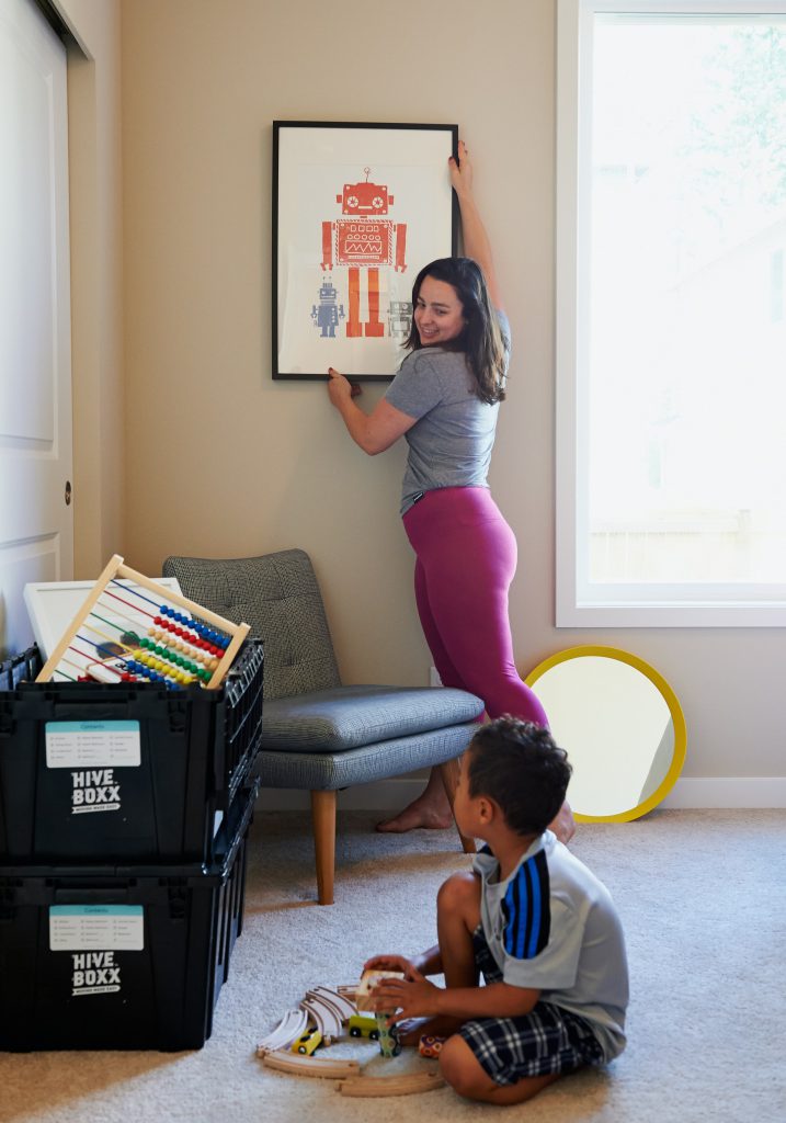  A imagem mostra uma mãe e um filho em uma sala. A mulher é branca, tem cabelo castanho-escuro liso, na altura dos ombros. Ela está em pé e ajusta o quadro de robôs na parede. O olhar dela está voltado para o menino, que está sentado no chão e brinca com alguns trenzinhos. Ao lado esquerdo do menino, há duas caixas plásticas pretas empilhadas. Na de cima, que está aberta, é possível ver um ábaco de madeira. 