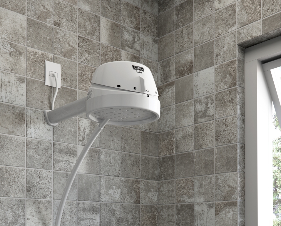 A imagem mostra uma ducha branca da Astra instalada em um banheiro com revestimento quadradinho em tom acinzentado.