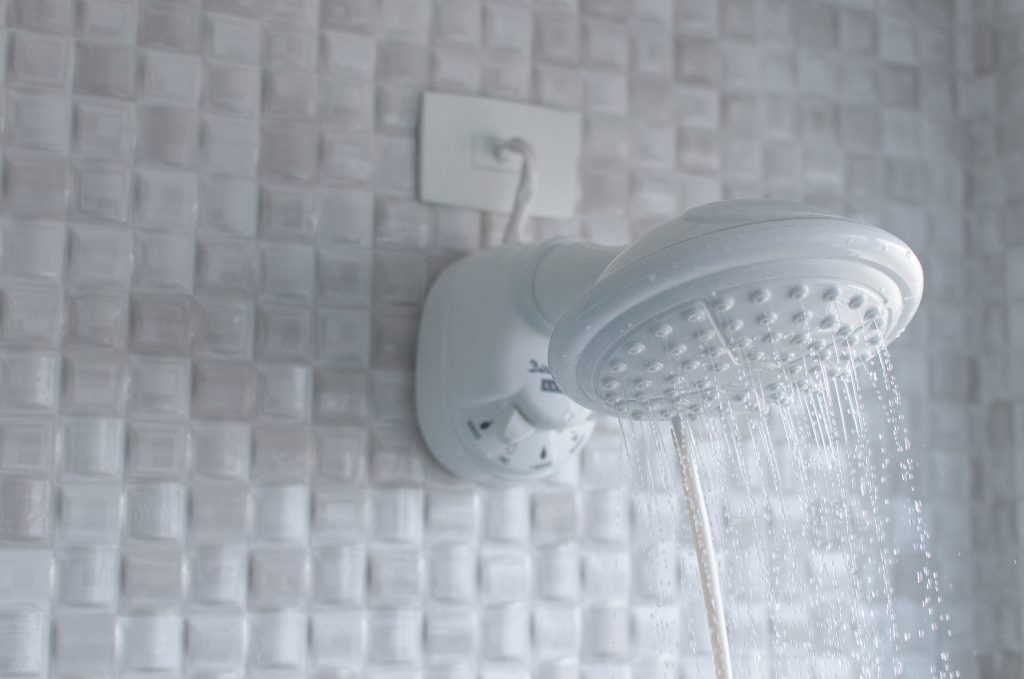 Um chuveiro branco da Astra está instalado em um banheiro com revestimento quadriculado branco. É possível ver a água saindo do chuveiro. 