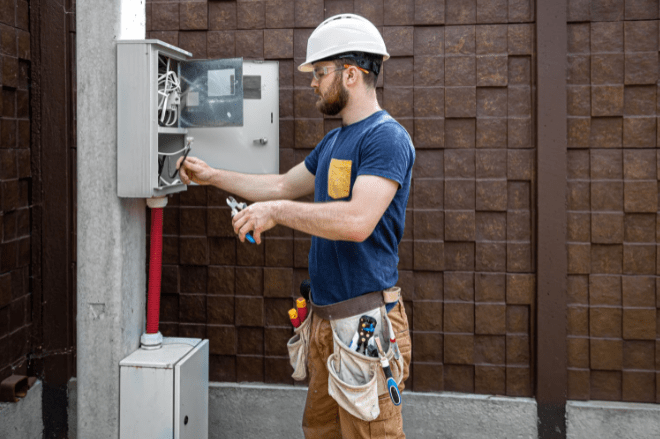 Um homem branco está mexendo em uma caixa de energia em um poste padrão. Ele usa equipamentos de segurança como capacete, óculos e luvas.