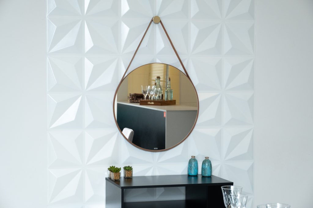 Um espelho redondo com alça está instalado em uma parede com revestimento 3D branco.
