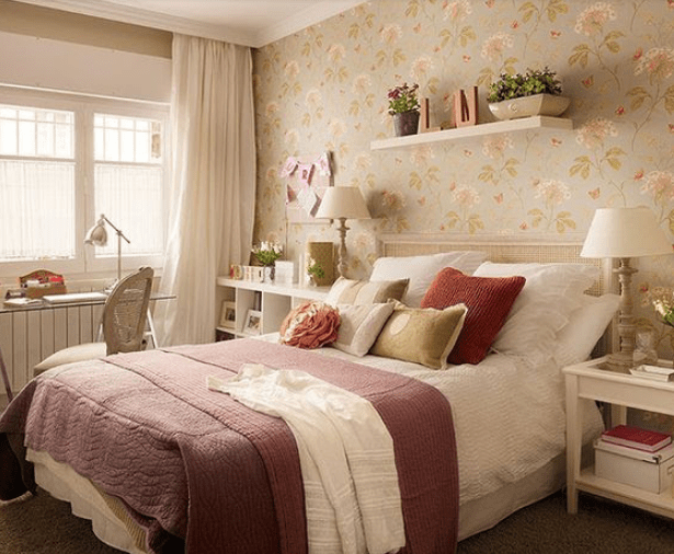 Um quarto com papel de parede florido aparece na imagem. Nele é possível ver uma cama de casal, uma escrivaninha, estante branca, prateleira branca e uma mesinha de cabeceira. 