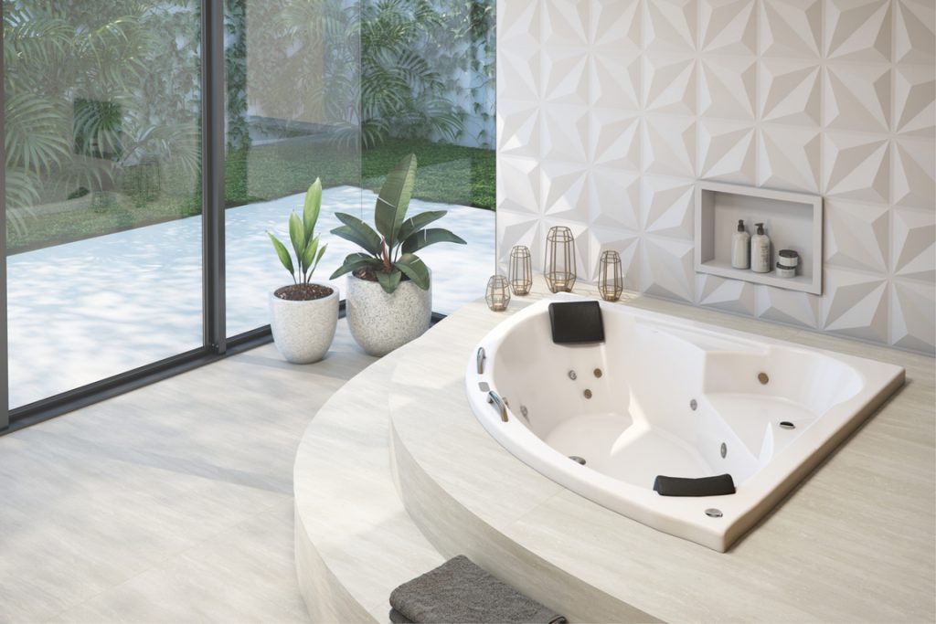 Uma banheira com formato de triângulo e espaço para duas pessoas está instalada em um chão de madeira em tom claro. Ao fundo há uma porta de vidro e dois vasos de planta. 