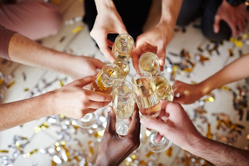 Sete mãos brindando com taças de champanhe com confetes dourados e prateados no chão ao fundo, como se fosse uma festa de ano novo.