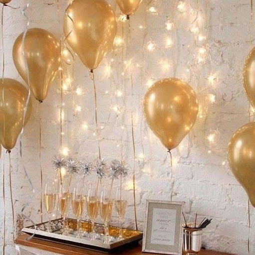 Sobre uma mesa de canto, uma bandeja com taças de champanhe, um quadro, e na parede estão coladas bexigas douradas e luzes pisca-piscas.
