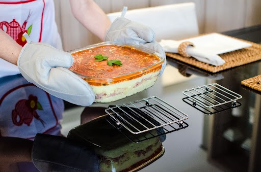 Uma pessoa de luvas segurando um refratário com comida e o apoiando sobre uma grelha que está em cima de uma mesa preta. 