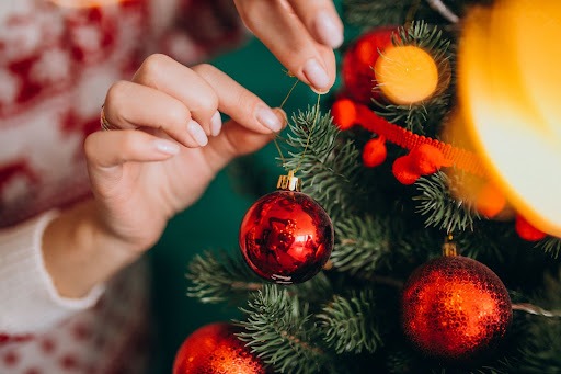 Uma mulher colocando uma bola de Natal vermelha com detalhes dourados em uma árvore com outras bolas e um enfeite vermelho.