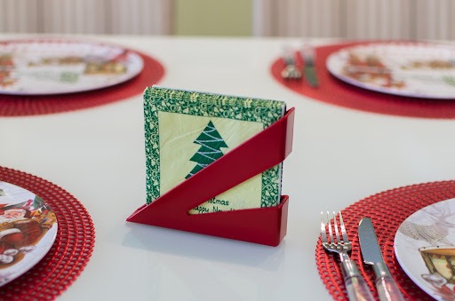 Mesa branca com porta-guardanapo vermelho da Astra ao centro. Ele está cheio de guardanapos verdes e brancos, que possuem desenho de árvore de Natal. Ao redor, estão quatro jogos americanos com pratos ilustrados de papai noel e talheres.
