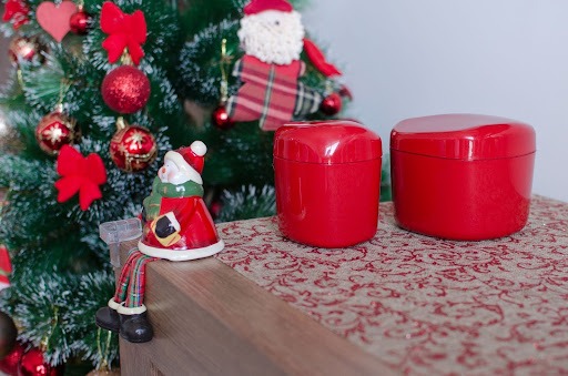 Ao fundo, uma árvore de Natal com enfeites em vermelho ao lado de um móvel com um boneco natalino apoiado em sua ponta. Um pouco mais atrás, estão um porta-guardanapo e um porta-cotonete vermelhos.