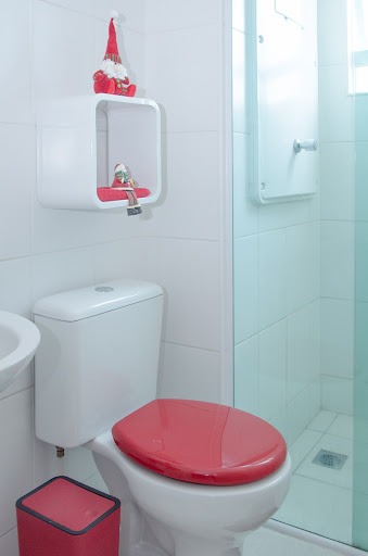 Um banheiro branco com lixeira e assento sanitário vermelhos e nicho quadrado na parede decorado com dois pequenos papais-noéis.

