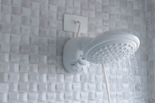 Um chuveiro branco da Astra está instalado em um banheiro com revestimento quadriculado branco. Na foto, ele aparece aberto e é possível ver a água saindo do aparelho.