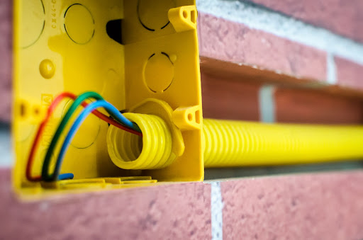 Uma caixa e tubo amarelos, com fios vermelho, verde e azul aparece embutida em uma parede de tijolos.