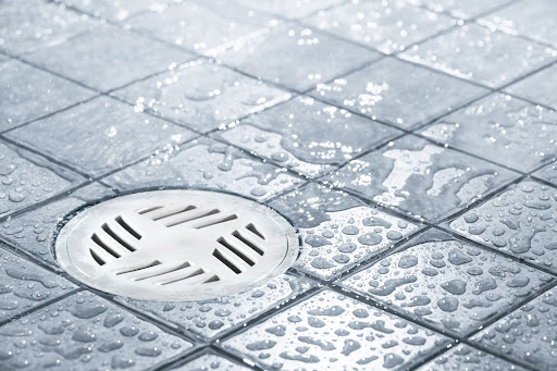 Um ralo de banheiro redondo branco da Astra aparece em um piso quadriculado cinza molhado. Esse é um dos tipos de ralos para banheiro.