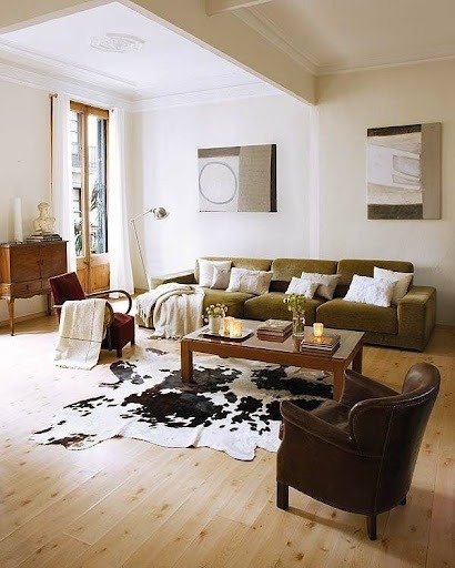 Uma sala de estar com um sofá, duas poltronas, uma mesa de centro, um móvel de madeira e um tapete de couro bovino. O piso é de madeira. 