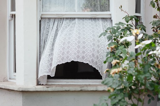 Uma janela aberta, com uma cortina branca esvoaçando ao vento. Há uma planta próxima à janela. 
