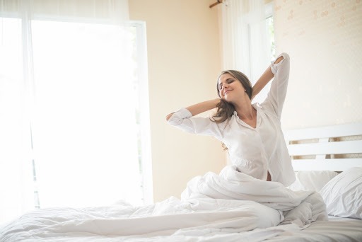 Uma mulher branca, vestindo pijama branco, se espreguiça ainda em cima da cama, cujos lençóis, cobertores e travesseiros também são brancos. Há uma porta de vidro ao fundo, coberta por uma cortina.