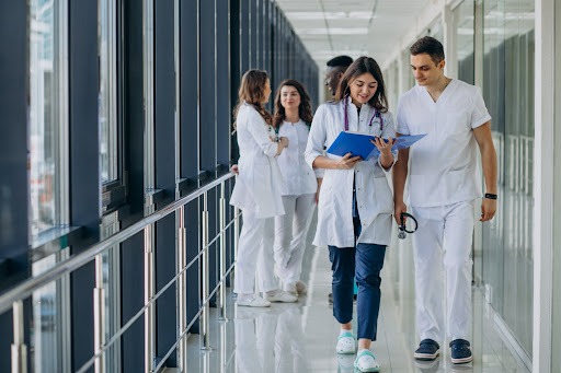  Um corredor de hospital. Vemos dois médicos brancos em primeiro plano, sendo um homem e uma mulher, andando e conversando. Os dois olham para um arquivo ou prontuário que está nas mãos da médica. Mais ao fundo, há um trio de médicos, sendo duas mulheres brancas e um homem negro, conversando. Eles estão parados.