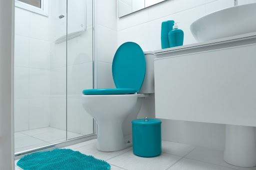Um banheiro com piso, revestimento, armário, cuba e vaso sanitário brancos. O assento e a tampa do vaso, a lixeira, o tapete e os acessórios em cima do armário são da cor spray, que se parece com um verde. Também é possível ver parte do box e parte do espelho.