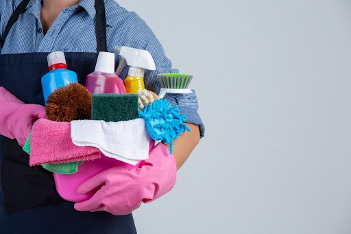 Uma pessoa usando luvas rosas e um avental segura um balde cheio de produtos de limpeza, como panos, escovas, esponjas e produtos químicos.
