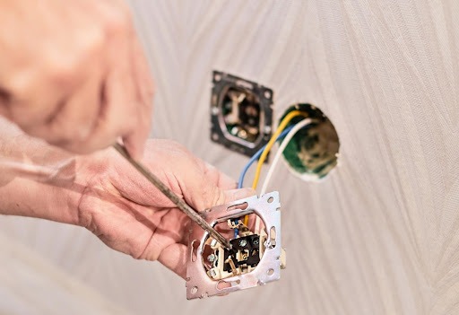 Uma pessoa branca usando uma chave para instalar uma tomada. Ao fundo, é possível ver outra tomada já instalada.